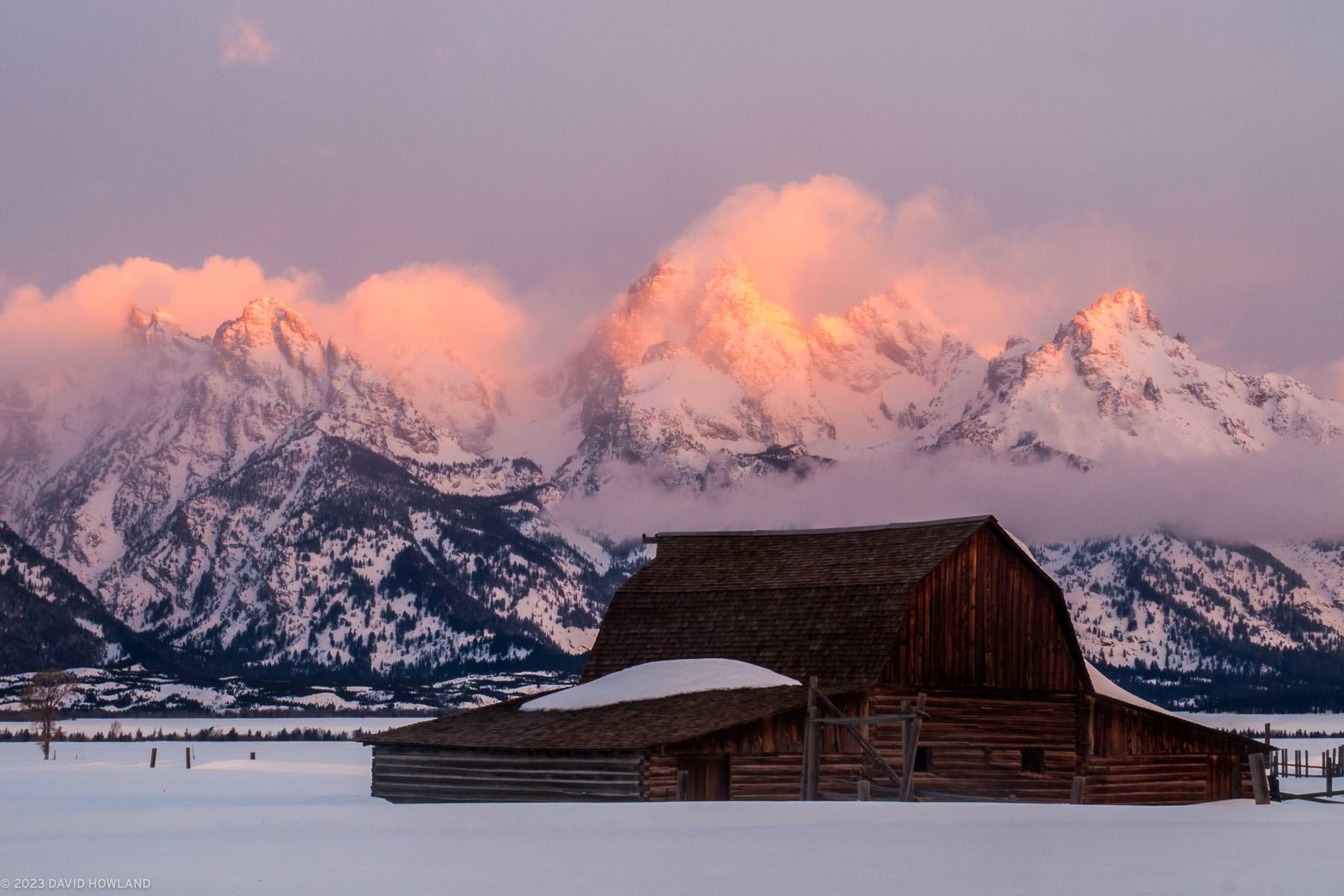 The rocky peaks of the Teton Range glow orange behind Moulton Barn in Grand Teton National Park.